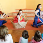 Drei Frauen spielen für Kinder; eine spielt Geige, eine sitzt in einem Korb und hält eine Schildkrötenpuppe, und eine spielt Akkordeon. Vier Kinder schauen vom Vordergrund aus zu.