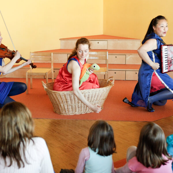 Drei Frauen spielen für Kinder; eine spielt Geige, eine sitzt in einem Korb und hält eine Schildkrötenpuppe, und eine spielt Akkordeon. Vier Kinder schauen vom Vordergrund aus zu.