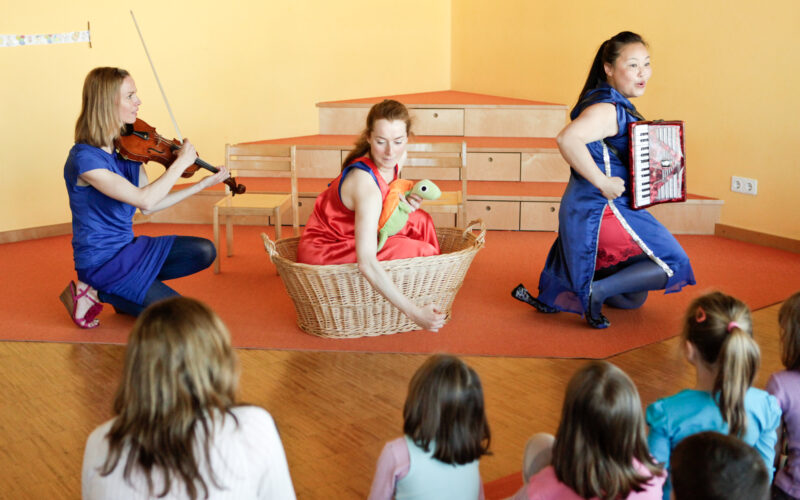Drei Frauen spielen für Kinder; eine spielt Geige, eine sitzt in einem Korb und hält eine Schildkrötenpuppe, und eine spielt Akkordeon. Vier Kinder schauen vom Vordergrund aus zu.