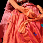 Eine Person in orange-roter Kleidung lehnt ihren Kopf an eine andere Person, die in ähnlich leuchtende Stoffe gehüllt ist, beide auf der Bühne vor einem dunklen Hintergrund.