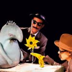 Zwei Personen mit Hüten und Sonnenbrillen interagieren mit einer blauen Marionette und einer gelben Blumenrequisite vor einem schwarzen Hintergrund.