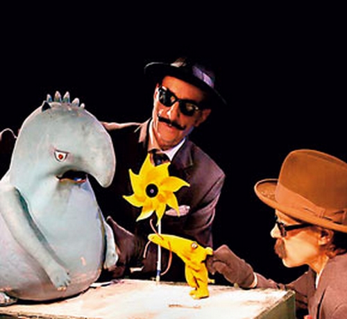 Zwei Personen mit Hüten und Sonnenbrillen interagieren mit einer blauen Marionette und einer gelben Blumenrequisite vor einem schwarzen Hintergrund.