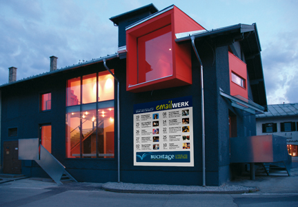 Modernes Gebäude mit großen, rot gerahmten Fenstern. Der Innenraum ist hell erleuchtet, und an der Außenwand neben dem Eingang hängt ein Veranstaltungsplakat.