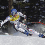 Ein Skifahrer im Rennanzug und mit Helm macht eine scharfe Kurve auf einer verschneiten Abfahrtspiste während eines alpinen Skirennens.