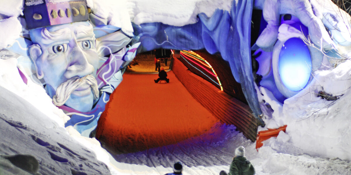 Zwei Personen auf Schlitten nähern sich einem Schneetunnel, der nachts mit einem bunten Gesicht und leuchtenden Lichtern geschmückt ist.