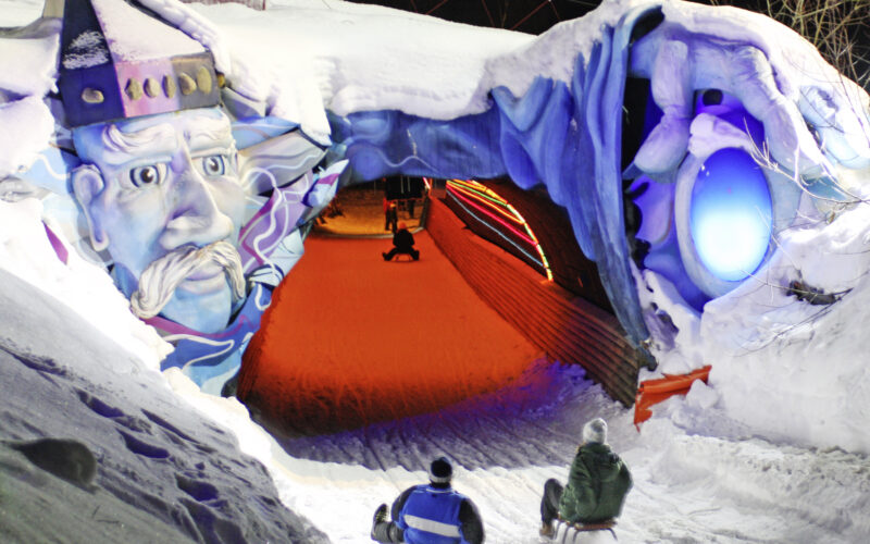 Zwei Personen auf Schlitten nähern sich einem Schneetunnel, der nachts mit einem bunten Gesicht und leuchtenden Lichtern geschmückt ist.