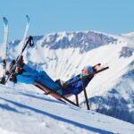 Eine Person in Skikleidung entspannt sich auf einem Liegestuhl an einem verschneiten Hang mit Bergen im Hintergrund und Skiern an der Seite.