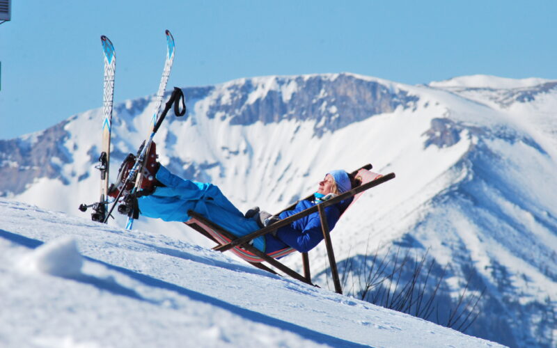 Eine Person in Skikleidung entspannt sich auf einem Liegestuhl an einem verschneiten Hang mit Bergen im Hintergrund und Skiern an der Seite.