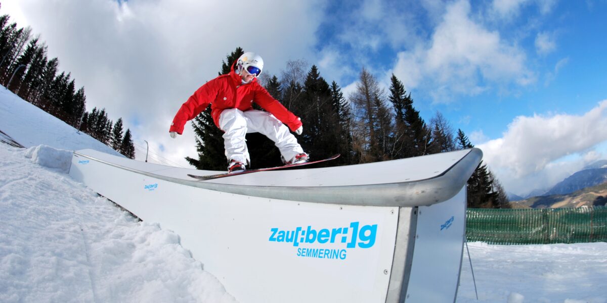 Ein Snowboarder in roter und weißer Ausrüstung gleitet auf einer Schiene in einem verschneiten Skigebiet mit Bäumen und Bergen im Hintergrund.