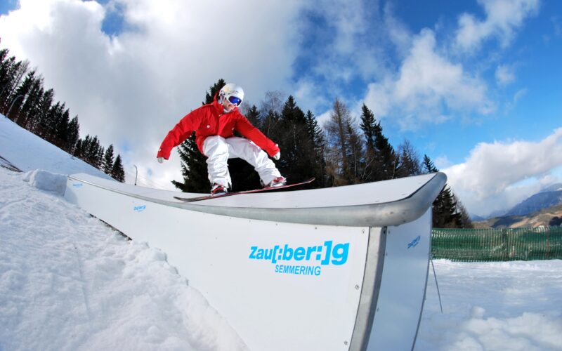 Ein Snowboarder in roter und weißer Ausrüstung gleitet auf einer Schiene in einem verschneiten Skigebiet mit Bäumen und Bergen im Hintergrund.