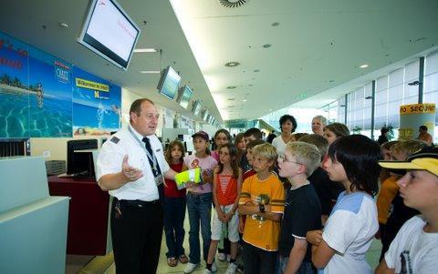 Ein uniformierter Flughafenmitarbeiter erklärt einer Gruppe von Kindern und Erwachsenen, die sich in der Nähe eines Abfertigungsschalters versammelt haben, die Sicherheitsmaßnahmen.