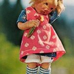 Eine Puppe mit unordentlichem blondem Haar, die ein rosa Kleid mit weißen Mustern und blau-gestreiften Socken trägt und eine kleine Blume in der Hand hält, steht im Freien.