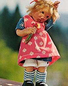 Eine Puppe mit unordentlichem blondem Haar, die ein rosa Kleid mit weißen Mustern und blau-gestreiften Socken trägt und eine kleine Blume in der Hand hält, steht im Freien.