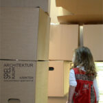 Ein junges Mädchen in einem roten Kleid steht im Freien und betrachtet ein großes Gebilde aus gestapelten Pappkartons, auf denen ein deutscher Text über Architektur steht.