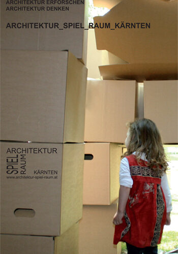 Ein junges Mädchen in einem roten Kleid steht im Freien und betrachtet ein großes Gebilde aus gestapelten Pappkartons, auf denen ein deutscher Text über Architektur steht.