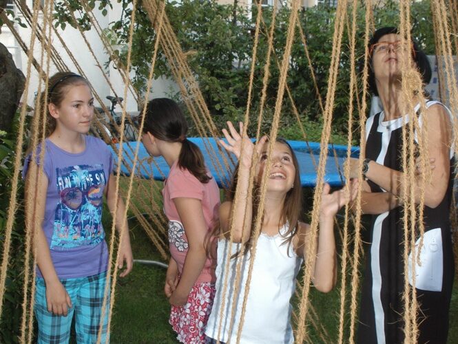 Vier Kinder und ein Erwachsener interagieren mit hängenden Seilen im Freien, mit einem Gebäude und Grünflächen im Hintergrund.