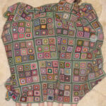 Eine Person liegt auf dem Rücken unter einer großen gehäkelten Decke mit einem Patchwork-Quadratmuster, wobei vier Füße am unteren Rand der Decke zu sehen sind.