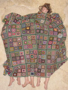 Eine Person liegt auf dem Rücken unter einer großen gehäkelten Decke mit einem Patchwork-Quadratmuster, wobei vier Füße am unteren Rand der Decke zu sehen sind.