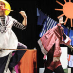 Zwei Darsteller auf der Bühne: einer im Matrosenoutfit und mit gelbem Hut, der einen Stock hält, der andere teilweise mit Kleidung bedeckt, mit einer Sonne und hängender Wäsche im Hintergrund.