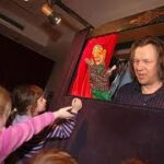 Ein Puppenspieler spielt mit zwei Puppen auf einer kleinen Bühne, während die Kinder zusehen und nach den Puppen greifen.