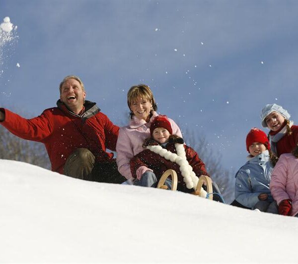 Eine Gruppe von Erwachsenen und Kindern sitzt auf einem verschneiten Hügel, einige auf einem Schlitten, in Winterkleidung unter einem klaren blauen Himmel.