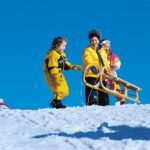 Drei Kinder in Winterkleidung stehen auf einem verschneiten Hügel mit einem Holzschlitten unter einem strahlend blauen Himmel.