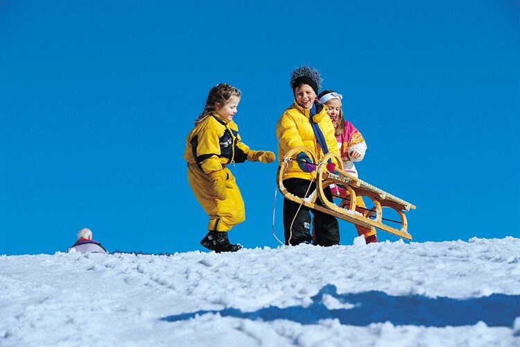 Drei Kinder in Winterkleidung stehen auf einem verschneiten Hügel mit einem Holzschlitten unter einem strahlend blauen Himmel.
