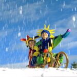 Zwei Kinder in farbenfrohen Kostümen mit Hüten sitzen auf einem Holzschlitten im Schnee, lächeln und spielen, während der Schnee vor einem strahlend blauen Himmel fällt.