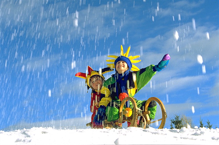 Zwei Kinder in farbenfrohen Kostümen mit Hüten sitzen auf einem Holzschlitten im Schnee, lächeln und spielen, während der Schnee vor einem strahlend blauen Himmel fällt.