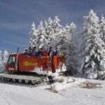 Eine Gruppe von Menschen steht auf einem roten Schneekatzenfahrzeug in einer verschneiten Landschaft, umgeben von schneebedeckten Bäumen unter einem klaren blauen Himmel.
