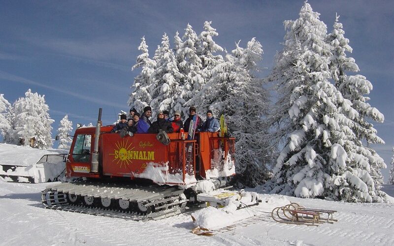 Eine Gruppe von Menschen steht auf einem roten Schneekatzenfahrzeug in einer verschneiten Landschaft, umgeben von schneebedeckten Bäumen unter einem klaren blauen Himmel.