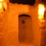 Ein Eingang aus festgefahrenem Schnee, der von warmen gelben Lichtern beleuchtet wird, führt zu einem weiteren schneebedeckten Bereich.