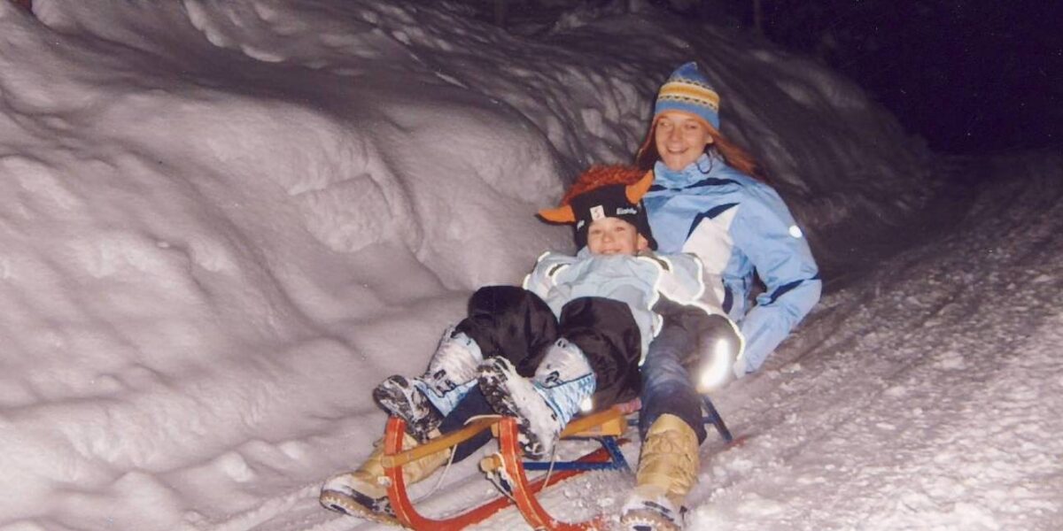 Ein Erwachsener und ein Kind in Winterkleidung schlittern nachts einen verschneiten Weg hinunter.