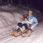 Ein Erwachsener und ein Kind in Winterkleidung schlittern nachts einen verschneiten Weg hinunter.