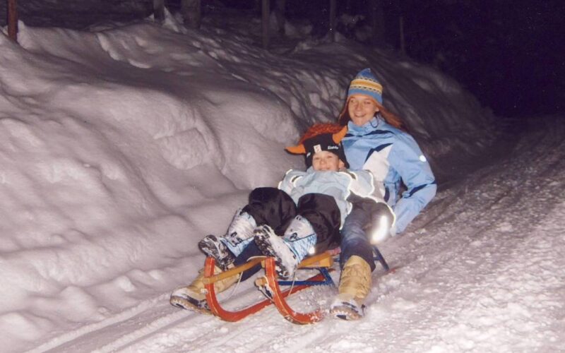 Ein Erwachsener und ein Kind in Winterkleidung schlittern nachts einen verschneiten Weg hinunter.