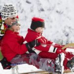 Ein Erwachsener schiebt zwei Kinder in Winterkleidung auf einem Holzschlitten in einer verschneiten Umgebung.