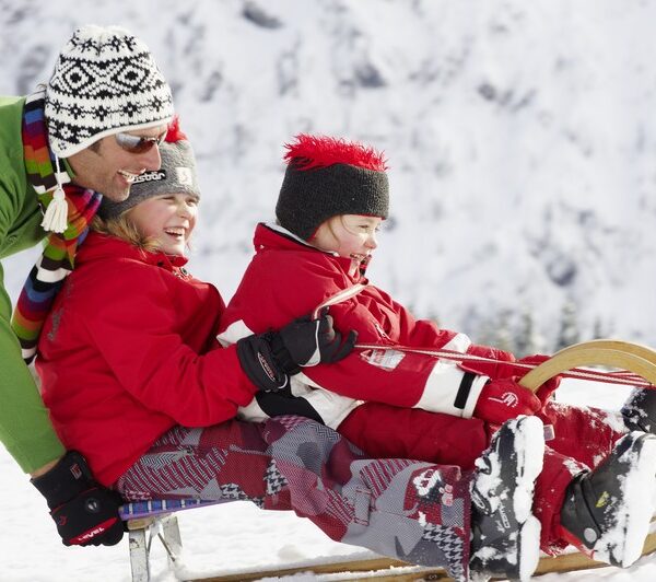 Ein Erwachsener schiebt zwei Kinder in Winterkleidung auf einem Holzschlitten in einer verschneiten Umgebung.