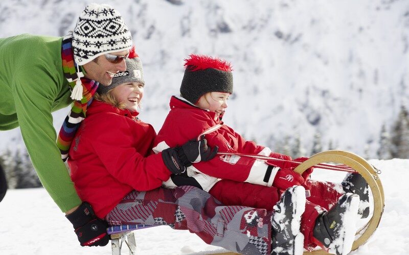 Ein Erwachsener schiebt zwei Kinder in Winterkleidung auf einem Holzschlitten in einer verschneiten Umgebung.