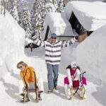 Zwei Frauen sitzen auf Schlitten, während ein Mann hinter ihnen mit erhobenen Armen auf einem verschneiten Weg mit hohen Schneewänden und schneebedeckten Häusern steht.