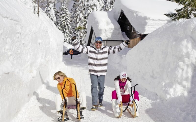 Zwei Frauen sitzen auf Schlitten, während ein Mann hinter ihnen mit erhobenen Armen auf einem verschneiten Weg mit hohen Schneewänden und schneebedeckten Häusern steht.