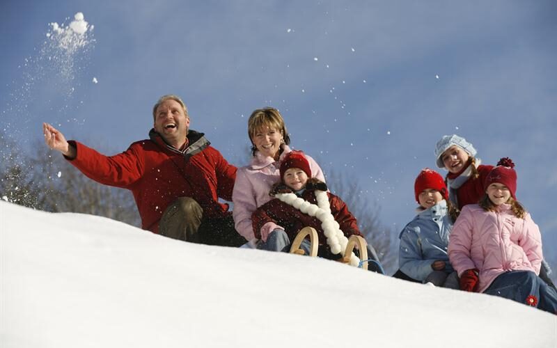 Sechs Personen, darunter Erwachsene und Kinder, sitzen in Winterkleidung auf einem verschneiten Hügel. Ein Erwachsener wirft Schnee, während ein Kind auf einem Schlitten sitzt. Der Himmel ist klar und blau.