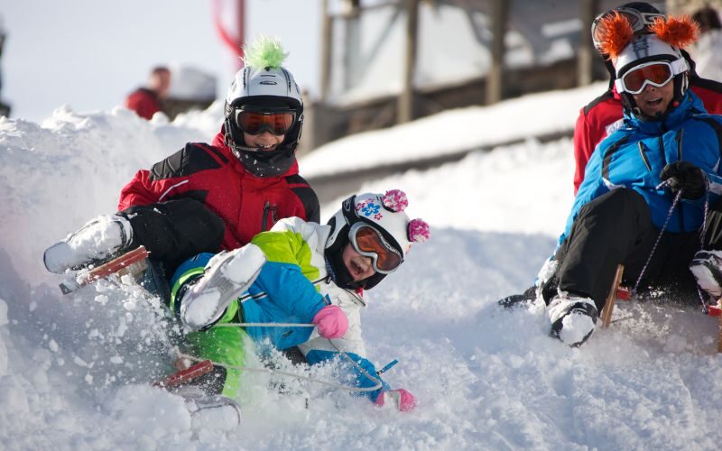 Drei Personen in bunten Winterklamotten und mit Helmen schlittern einen verschneiten Hügel hinunter und werfen dabei Schnee auf.