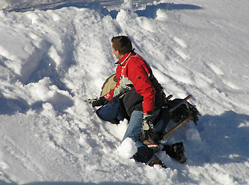 Eine Person in einer roten Jacke sitzt auf einem Schlitten am Fuße eines verschneiten Hangs, der teilweise unter Schnee begraben ist.