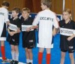 Eine Gruppe von Kindern, die schwarze und weiße Fußballtrikots tragen, steht in einer Reihe auf einem blauen Hallensportplatz.