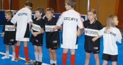 Eine Gruppe von Kindern, die schwarze und weiße Fußballtrikots tragen, steht in einer Reihe auf einem blauen Hallensportplatz.