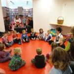 Eine Gruppe kleiner Kinder sitzt in einem Kreis auf einem roten Boden und hört einem Erwachsenen zu, während mehrere andere Erwachsene von nahegelegenen Sitzplätzen in einer Bibliothek oder einem Klassenzimmer aus zusehen.