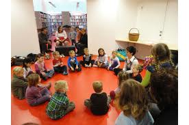 Eine Gruppe kleiner Kinder sitzt in einem Kreis auf einem roten Boden und hört einem Erwachsenen zu, während mehrere andere Erwachsene von nahegelegenen Sitzplätzen in einer Bibliothek oder einem Klassenzimmer aus zusehen.