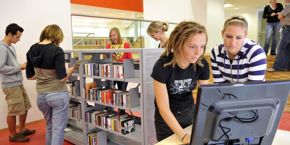 Mehrere junge Leute benutzen Computer und stöbern in den Bücherregalen einer modernen, hell erleuchteten Bibliothek.
