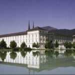 Ein großes weißes Gebäude mit zwei Türmen spiegelt sich in einem ruhigen See, mit Bäumen und Bergen im Hintergrund unter einem klaren Himmel.