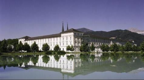 Ein großes weißes Gebäude mit zwei Türmen spiegelt sich in einem ruhigen See, mit Bäumen und Bergen im Hintergrund unter einem klaren Himmel.